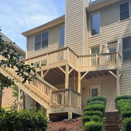 Two-story beige townhouse with a wooden deck, stairs, and landscaped shrubs on a sloped lot