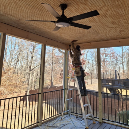 Person on a ladder installing a ceiling fan on a covered porch with wooded yard beyond.