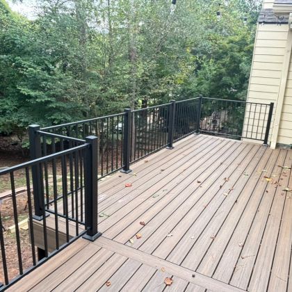 Wooden deck with black railings beside a beige house, overlooking a wooded backyard.
