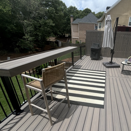 Wooden deck with striped rug, chair, grill, and umbrella overlooking a wooded creek and nearby houses