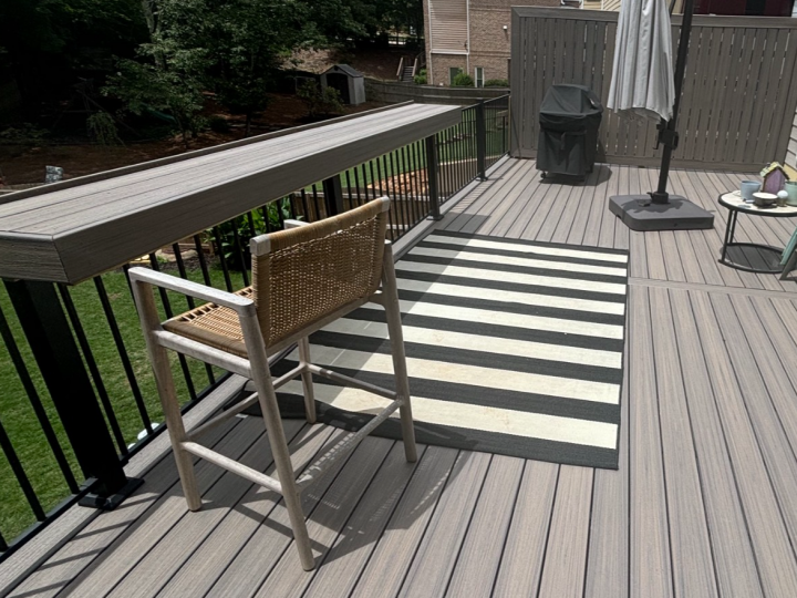 Wooden deck with striped outdoor rug, chair, railing, and patio umbrella on a backyard patio.