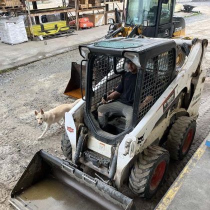 Worker in a white skid steer loader beside a small dog on a construction site.