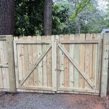 Wooden double gate in a privacy fence, with trees and a gravel drive behind it.