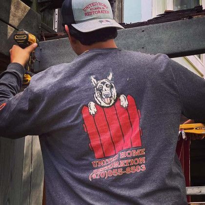 Worker using a drill on a wooden frame, wearing a gray cap and T-shirt with a dog logo on the back.