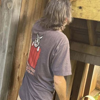 Person in a gray shirt with a red graphic standing by wooden beams outdoors.