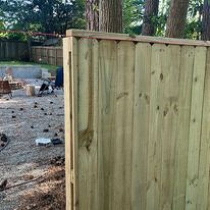 Wooden fence panel beside a gravel yard with scattered debris and trees in the background