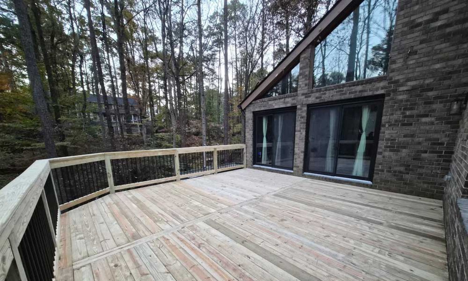 Wooden deck with railing beside a modern stone cabin, overlooking a wooded backyard.