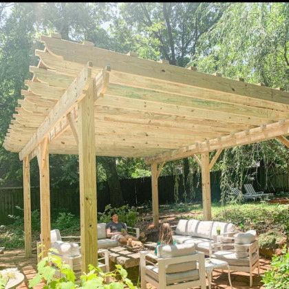 Wooden pergola over an outdoor patio with white sofas and chairs in a wooded backyard