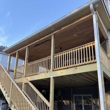Wooden two-story house porch with railings and stairs, viewed from below against a blue sky