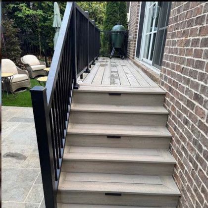Back steps with black railing leading to a wooden deck beside a brick house and patio area