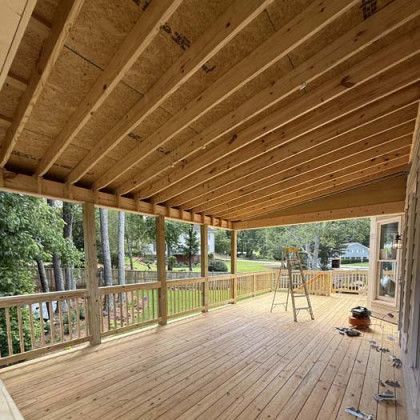 Wooden covered deck with railing, overlooking trees and a yard, with a ladder and grill nearby.