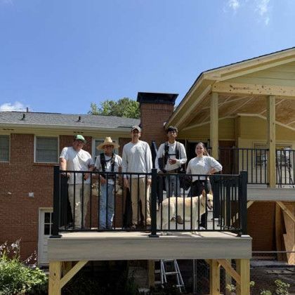 Five people stand on a black-railed porch of a brick house under a clear blue sky.