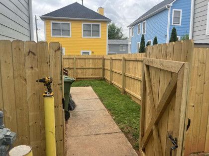Narrow fenced backyard walkway with concrete path, grass strip, open gate, and yellow and blue houses beyond.