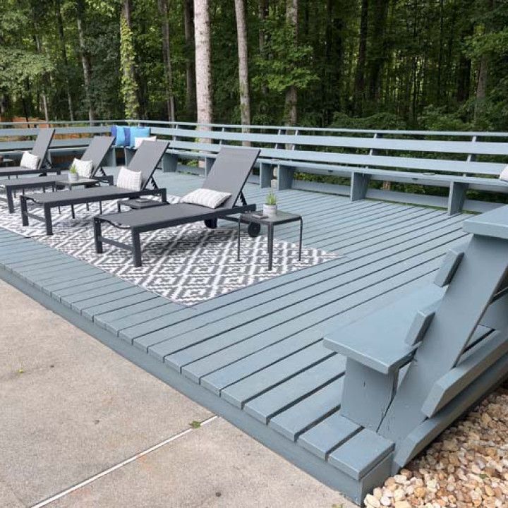 Gray outdoor deck with lounge chairs, patterned rug, and railing in a wooded setting