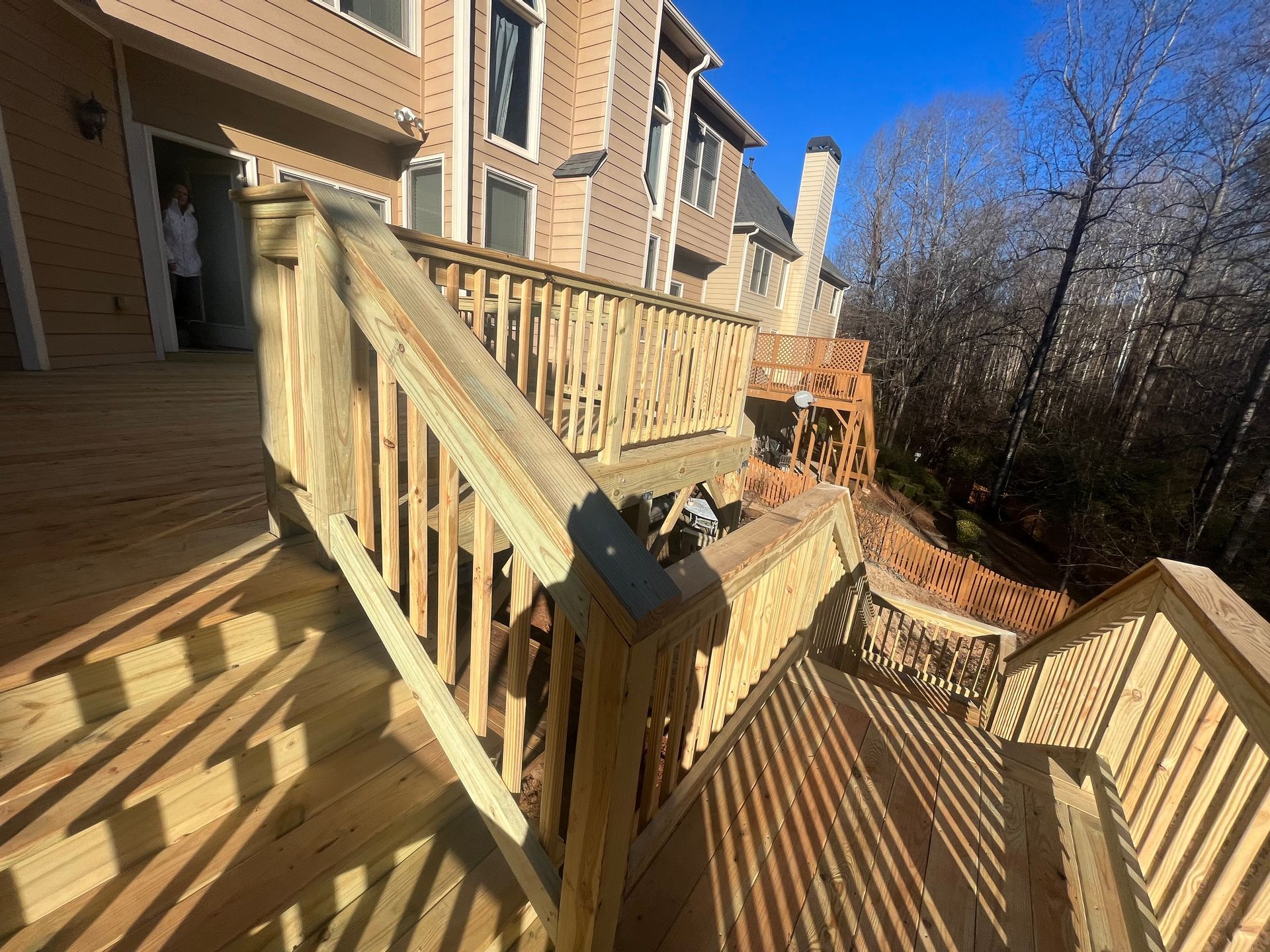Wooden deck and stairs beside a brick house, with sunlit railings and leafless trees nearby