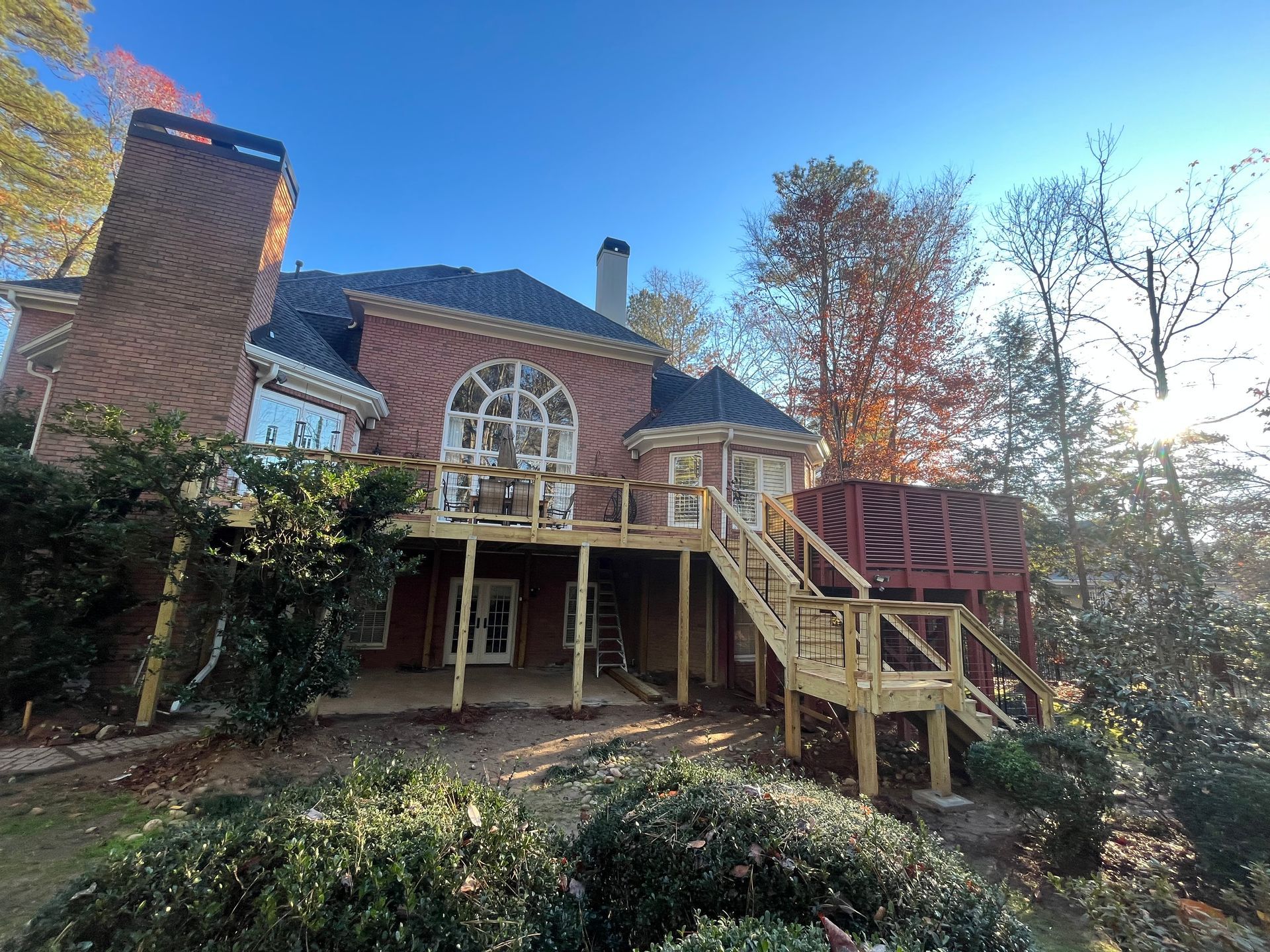Brick house with a large arched window, wooden deck and stairs, surrounded by trees in bright sunlight