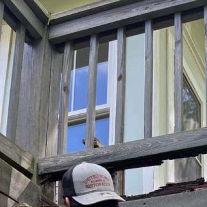 Person in a hard hat below a wooden railing, looking up at a window and weathered house siding.