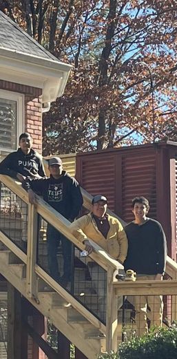 Four people standing on outdoor wooden steps beside a house, with autumn trees in the background.