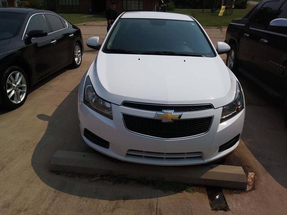 White Chevrolet sedan parked between two dark cars in a lot, with a concrete wheel stop in front.