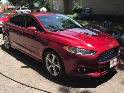 Red sedan parked on a residential street in front of houses, viewed from the front-left side.