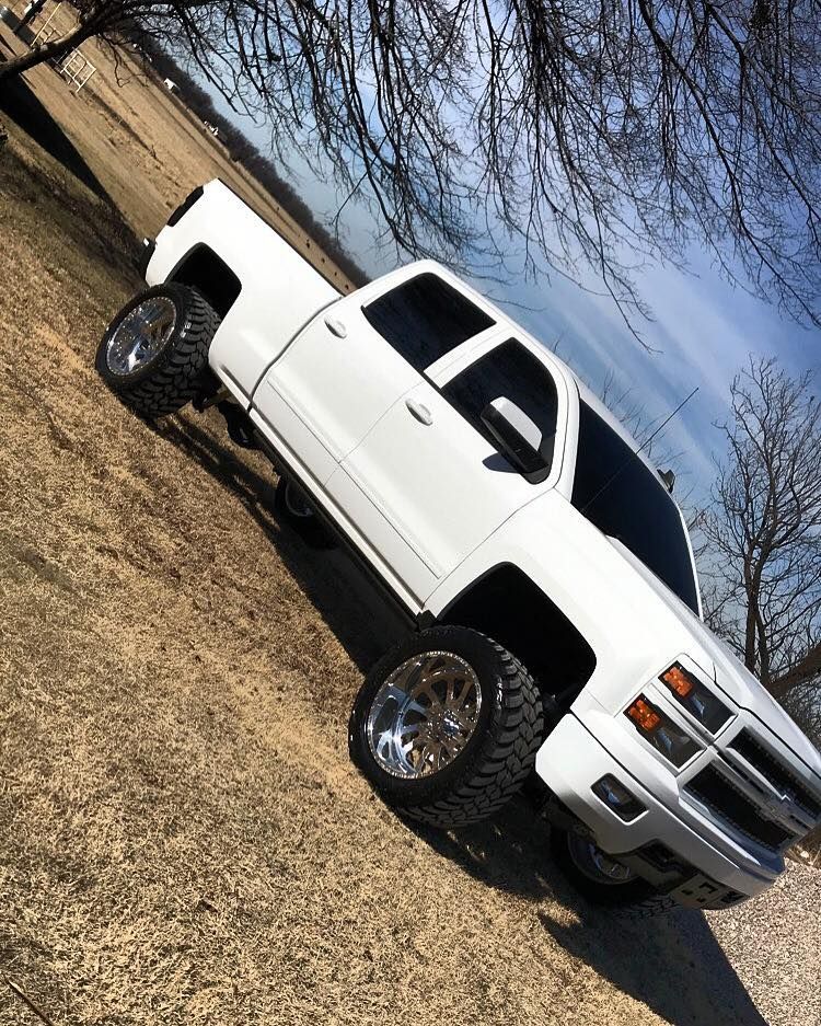 White lifted pickup truck parked on a grassy roadside near a wooden fence, tilted view