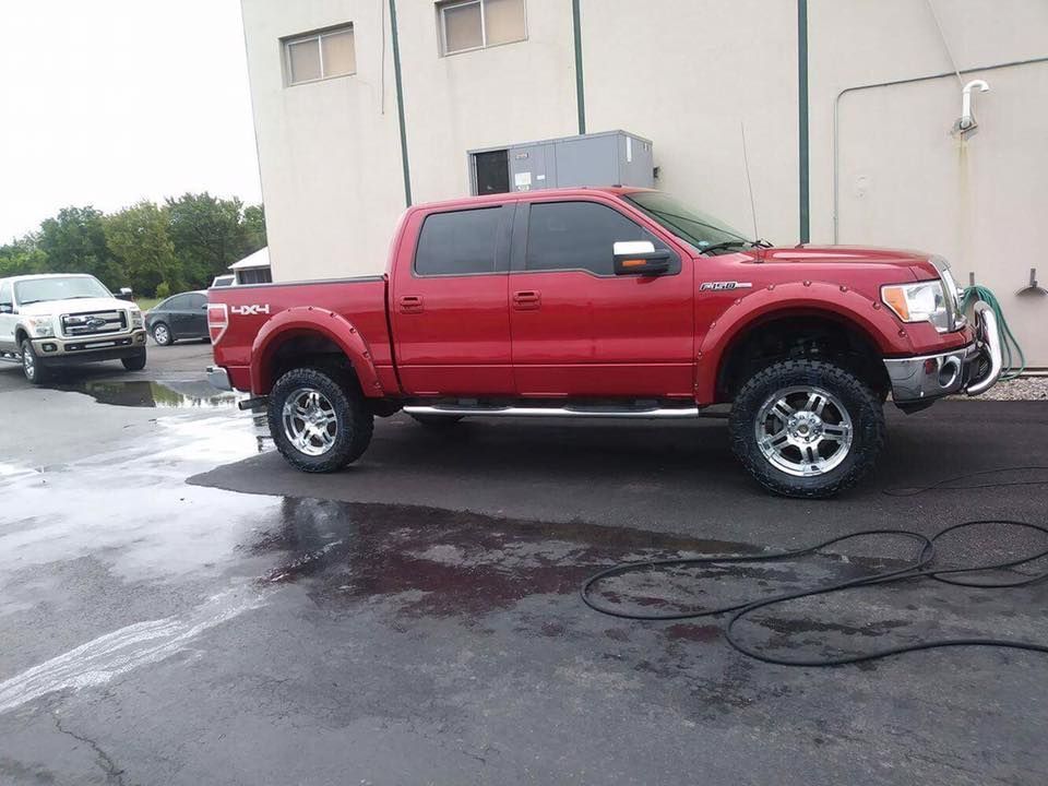 Red lifted pickup truck parked on wet pavement beside a white building, with a dark SUV in the background.
