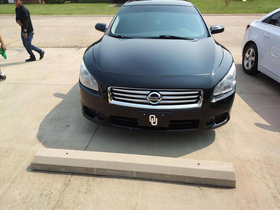Black Nissan sedan parked head-on in a driveway with a concrete curb stop in front