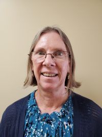 Smiling person wearing glasses, blue patterned top, and navy cardigan against a beige wall