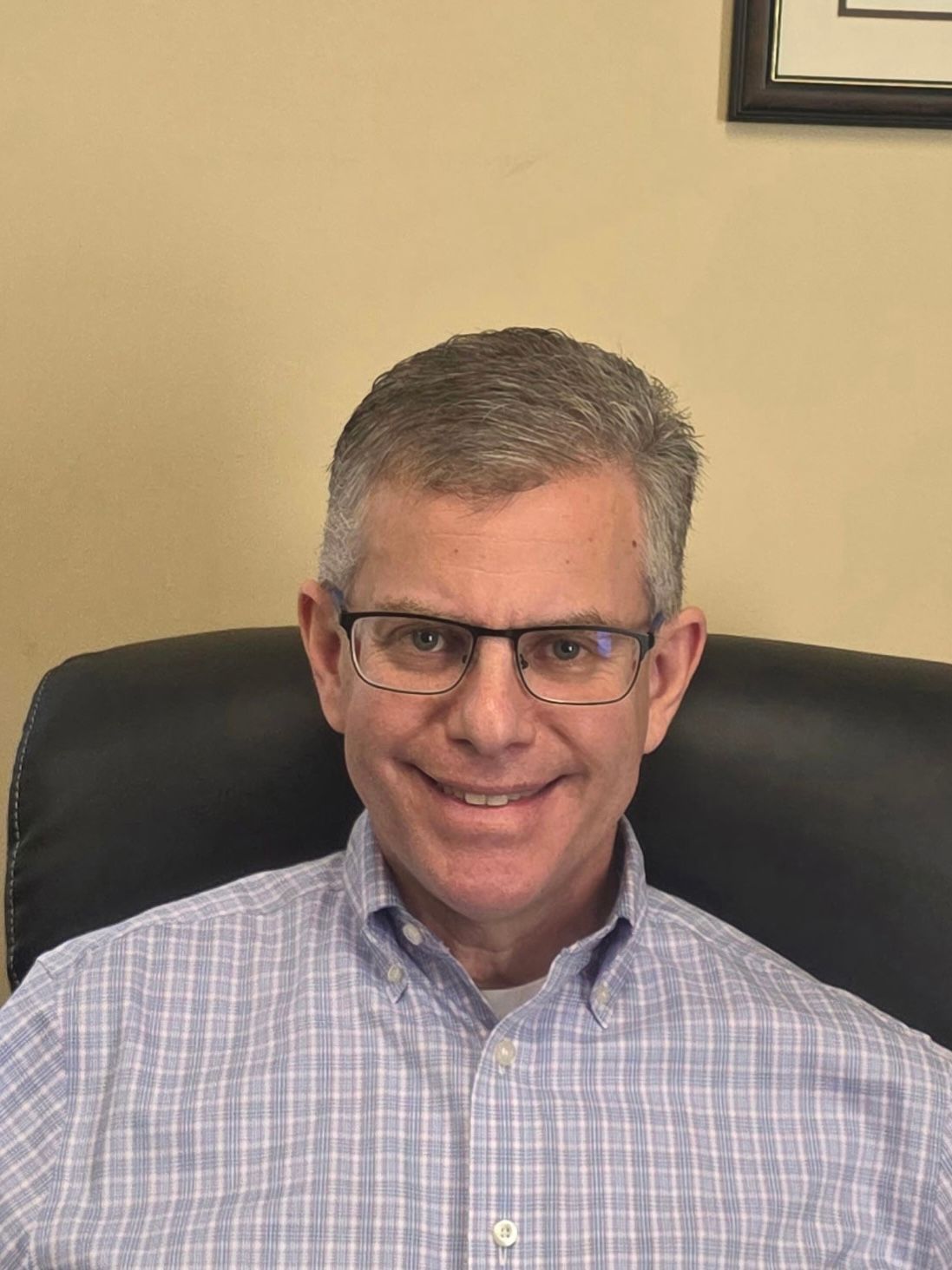 Smiling man wearing glasses seated in an office chair against a beige wall