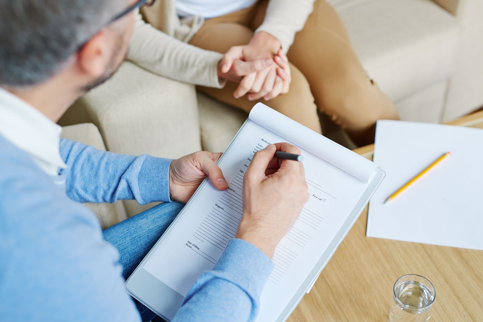 Person takes notes during a counseling session while two others sit on a couch nearby.