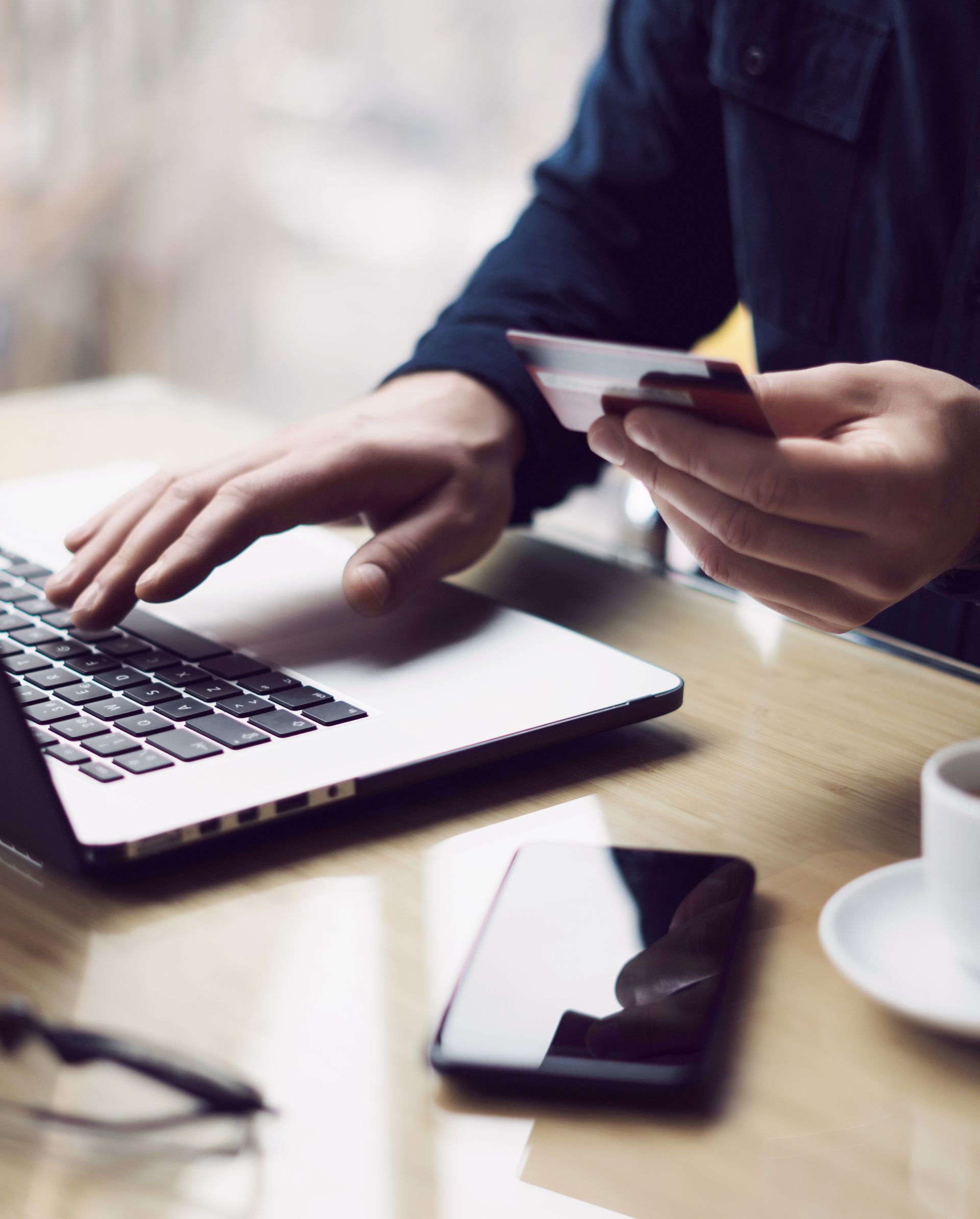 Person using a laptop and holding a credit card at a desk with a smartphone and coffee cup