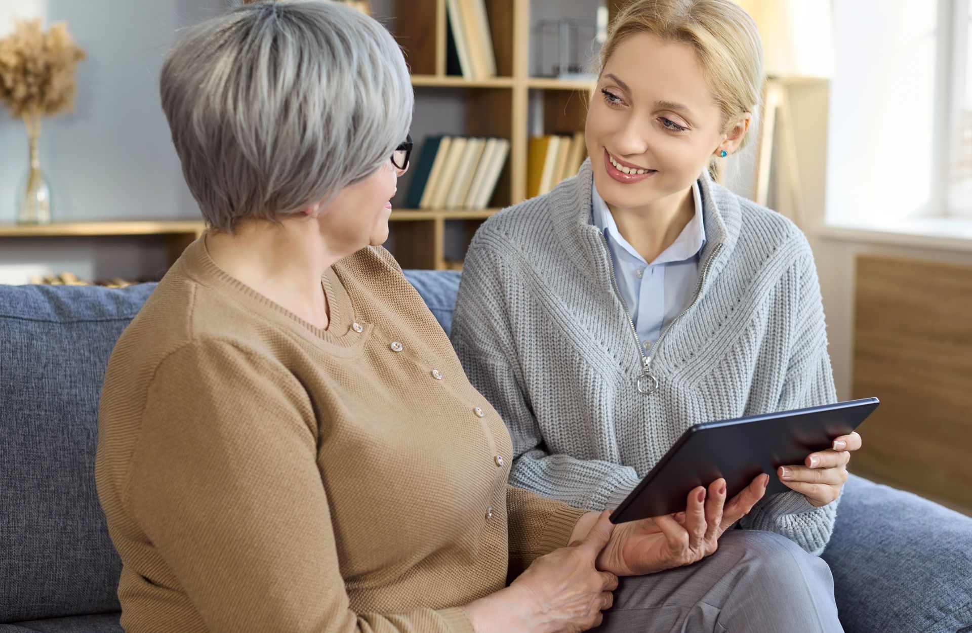Two people talking on a couch, one holding a tablet, in a bright living room