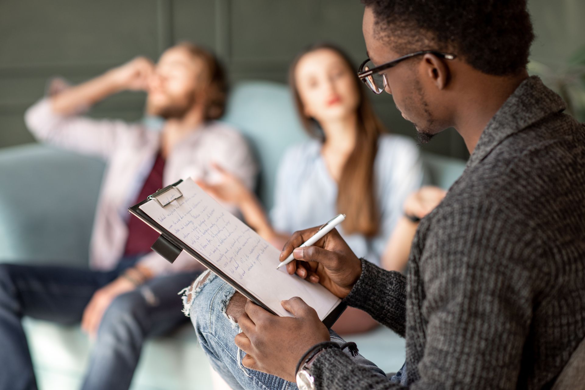 Therapist taking notes on a clipboard while speaking with a couple on a couch