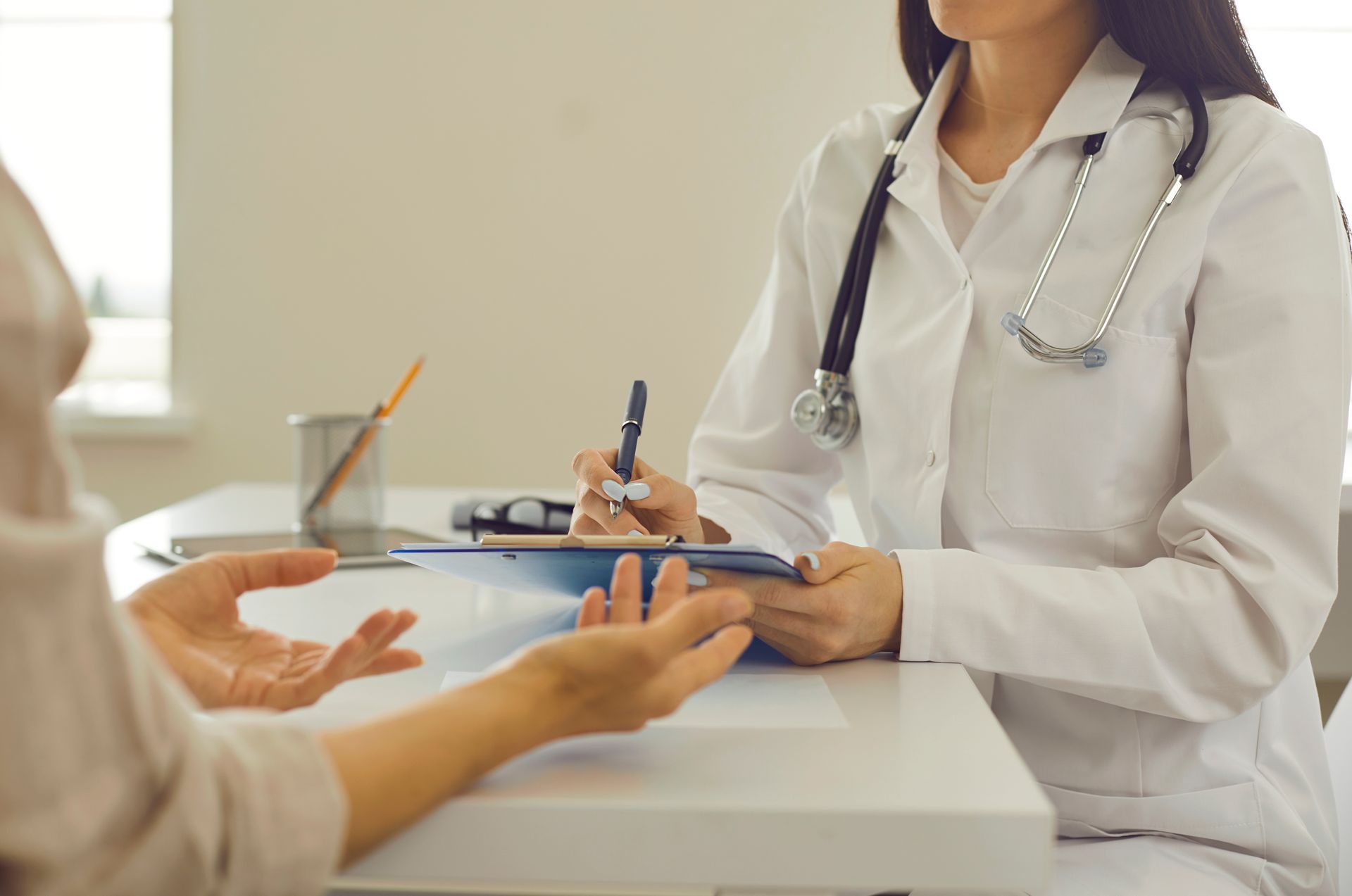 Doctor consulting with a patient across a desk, holding a clipboard in a clinic office.