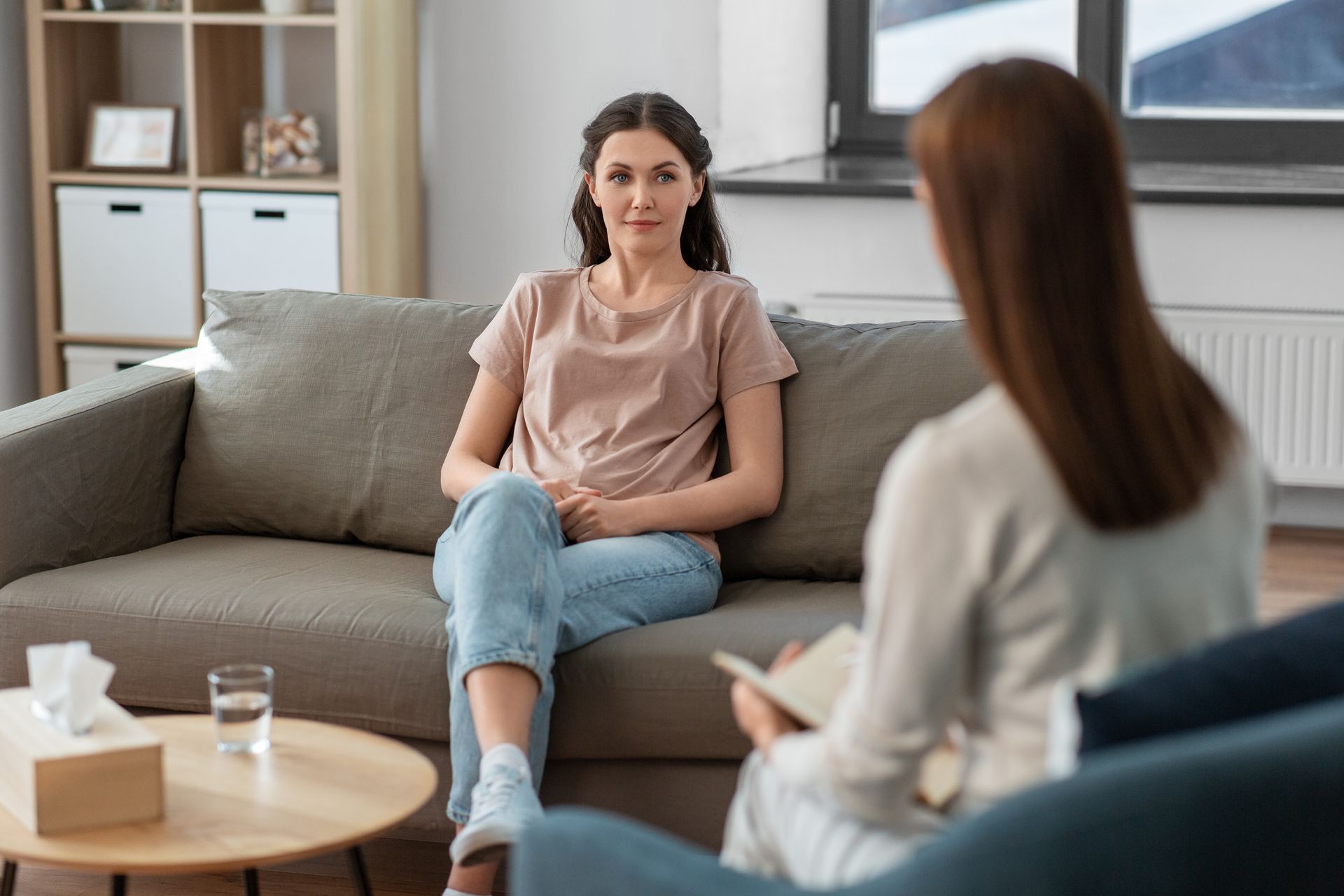 Two people talking in a living room, one seated on a gray sofa and another in the foreground.