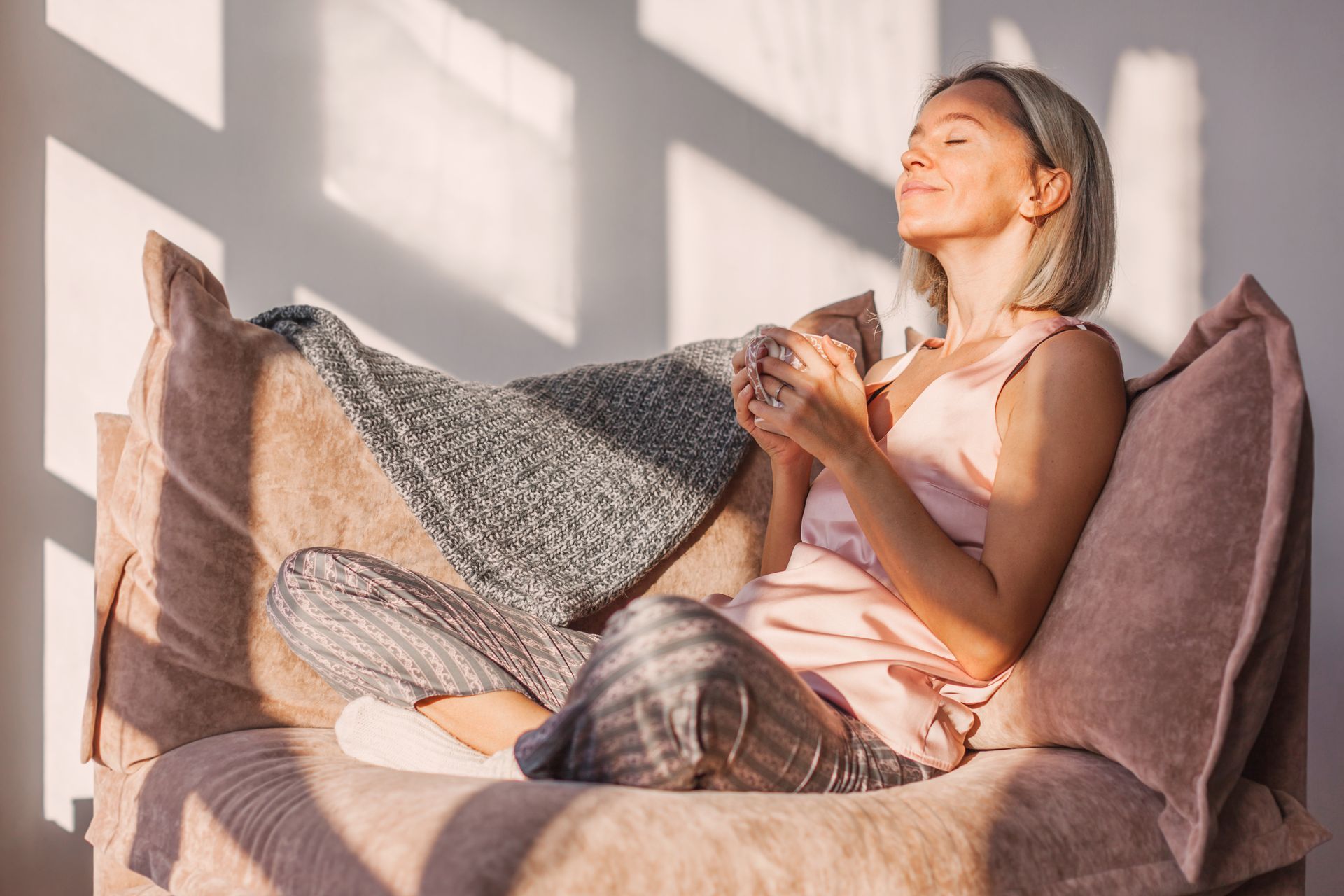 Woman relaxing on a couch by a sunlit window, wrapped in a blanket and holding a mug