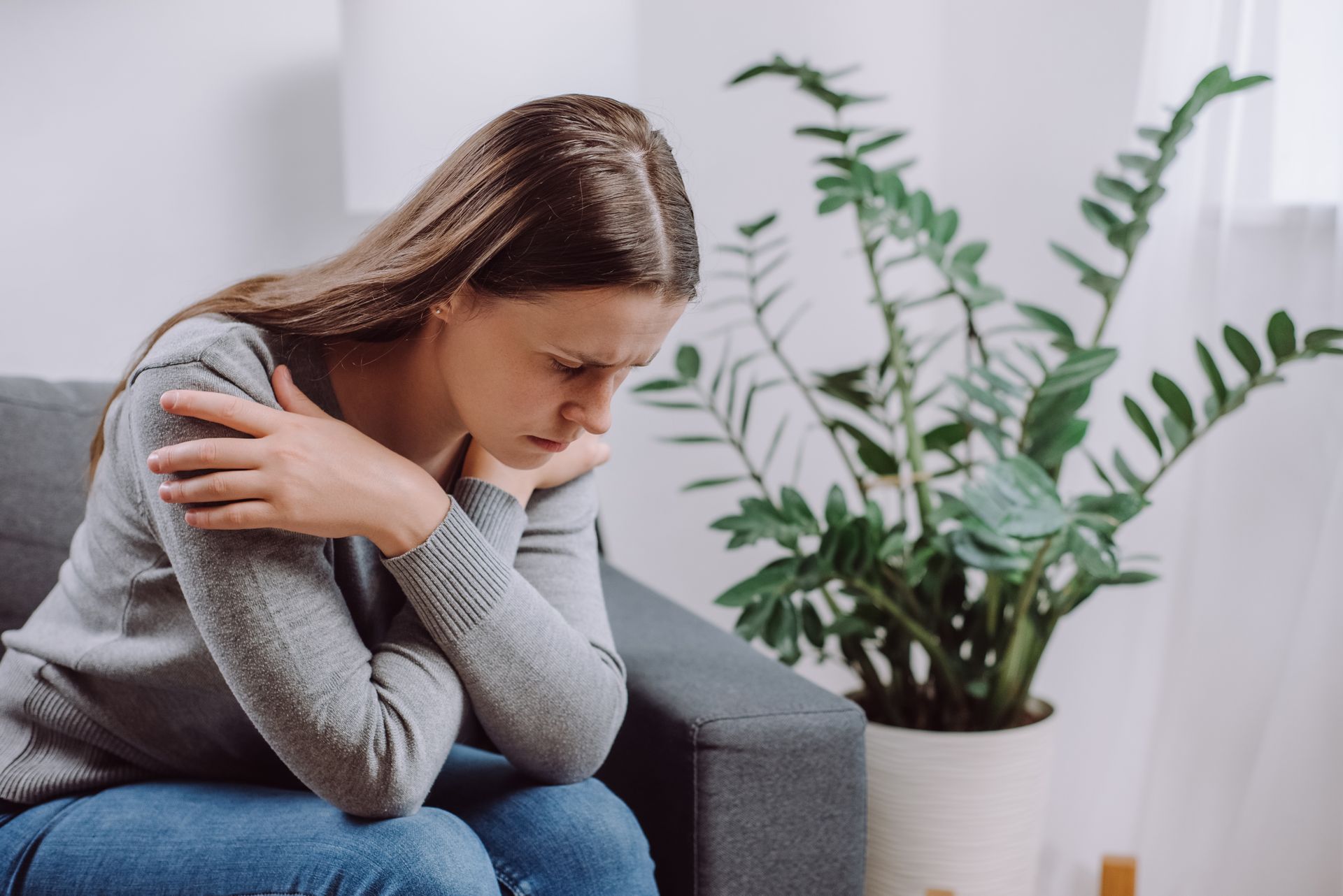 Person sitting on a couch, hugging themselves and looking down in a bright room with a potted plant.