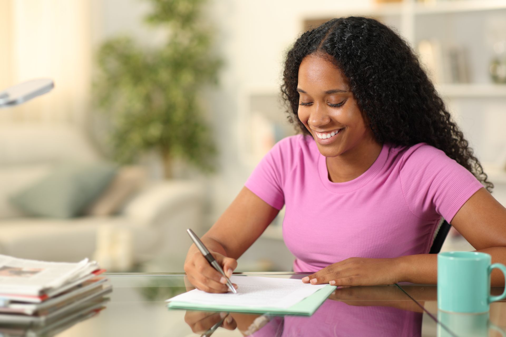Woman smiling and writing at a glass table with a mug, stack of papers, and laptop in a bright living room