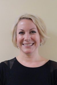 Smiling person with short blonde hair wearing a black top against a beige background