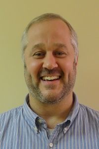 Smiling man in a blue striped shirt against a plain beige background