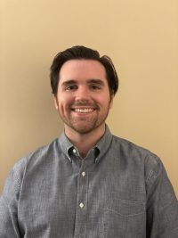 Smiling man in a gray button-up shirt standing against a plain beige wall