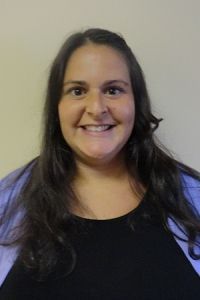 Smiling person with long dark hair wearing a black top and lavender sleeves against a plain wall