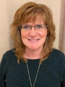 Portrait of a smiling woman wearing glasses and a dark green top against a beige background