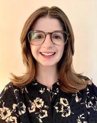 Smiling person with shoulder-length hair and glasses wearing a black floral blouse against a light background