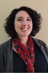 Smiling person with curly dark hair wearing a red scarf and gray cardigan against a plain wall