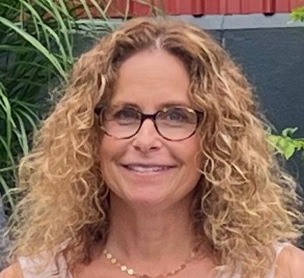 Smiling person with curly hair and glasses in a rust-colored shirt, standing before greenery.