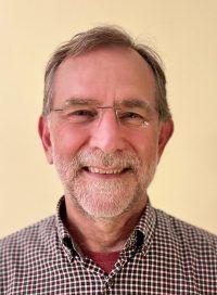 Smiling man with glasses and gray beard in a checkered shirt against a beige background