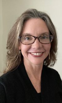 Smiling woman with glasses and long gray hair, wearing a black top against a plain background