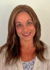 Smiling woman with long brown hair wearing a light cardigan and patterned top against a plain background