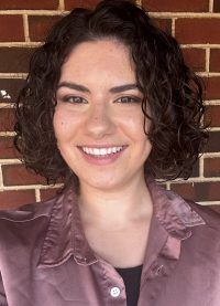 Smiling person with curly dark hair in a mauve shirt, standing in front of a brick wall.